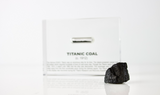 Close-up pieces of coal from the Titanic, contained in a small glass ampule and embedded in clear acrylic. The coal is dark and granular, offering a tangible link to the historic RMS Titanic.