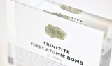 Close-up of a fragment of Trinitite, a material formed from the first atomic bomb explosion, embedded in clear acrylic. The piece has a greenish tint and a rough, uneven surface with visible texture, reflecting its unique origin from the historic Trinity test.