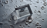 Close-up piece of V-2 rocket shrapnel embedded in clear acrylic. The fragment is metallic and jagged, with a distinctive shape that highlights its history from World War II rocket warfare.