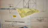 Close-up of a piece of fabric from the Wright Flyer, encapsulated in clear acrylic. The fabric shows a slightly worn texture with visible stitching, showcasing the early aviation history of the Wright brothers' first successful aircraft.