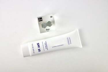 Zinc element encased in a clear acrylic cube, featuring its name, atomic number 30, and properties as displayed on the periodic table. Ideal for educational and collector use. Sunscreen, as a mass use being zinc oxide, sits next to the cube.