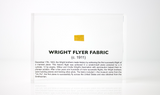 Close-up piece of fabric from the Wright Flyer, encapsulated in clear acrylic. The fabric shows a slightly worn texture with visible stitching, showcasing the early aviation history of the Wright brothers' first successful aircraft.