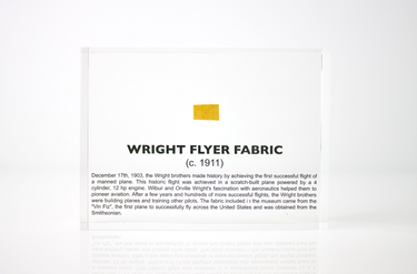 Close-up piece of fabric from the Wright Flyer, encapsulated in clear acrylic. The fabric shows a slightly worn texture with visible stitching, showcasing the early aviation history of the Wright brothers' first successful aircraft.