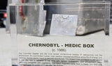 Clear acrylic display case featuring a historical fragment of a Chernobyl medic box, offering an educational presentation for collectors and those interested in nuclear history.