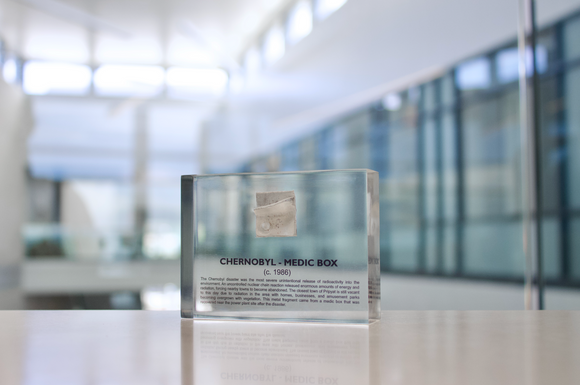 Clear acrylic display case featuring a historical fragment of a Chernobyl medic box, offering an educational presentation for collectors and those interested in nuclear history.