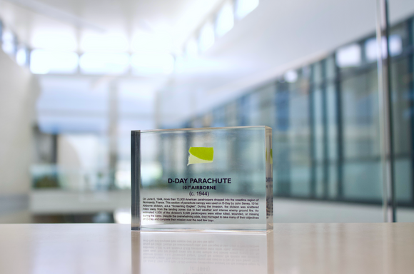 Clear acrylic display case featuring a historical fragment of a D-Day parachute, offering an educational presentation for collectors and those interested in World War II history.