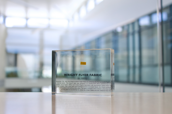 Close-up piece of fabric from the Wright Flyer, encapsulated in clear acrylic. The fabric shows a slightly worn texture with visible stitching, showcasing the early aviation history of the Wright brothers' first successful aircraft.