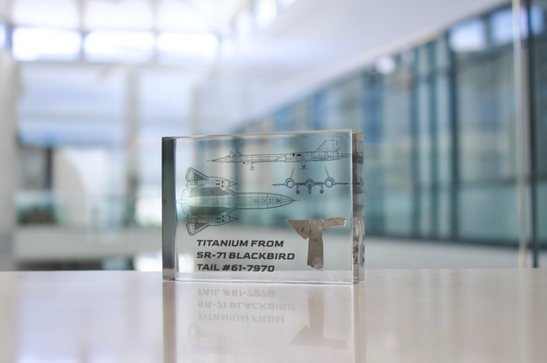 Close-up piece of SR-71 Blackbird titanium embedded in clear acrylic. The fragment is metallic and jagged, which highlights its history from the high-speed reconnaissance aircraft. A blueprint of the SR-71 aircraft is visible in the display.