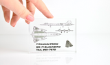 Close-up piece of SR-71 Blackbird titanium embedded in clear acrylic. The fragment is metallic and jagged, which highlights its history from the high-speed reconnaissance aircraft. A blueprint of the SR-71 aircraft is visible in the display.