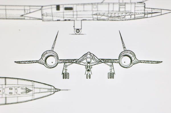 Close-up piece of SR-71 Blackbird titanium embedded in clear acrylic. The fragment is metallic and jagged, which highlights its history from the high-speed reconnaissance aircraft. A blueprint of the SR-71 aircraft is visible in the display.