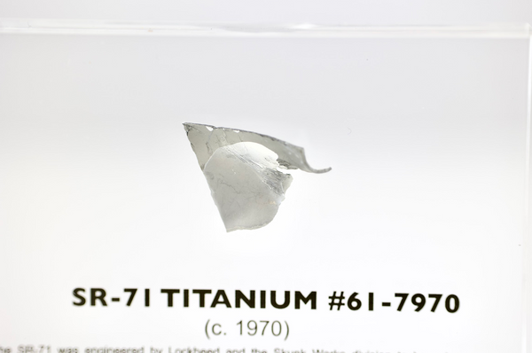 Close-up piece of SR-71 Blackbird titanium embedded in clear acrylic. The fragment is metallic and jagged, which highlights its history from the high-speed reconnaissance aircraft.