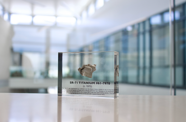 Close-up piece of SR-71 Blackbird titanium embedded in clear acrylic. The fragment is metallic and jagged, which highlights its history from the high-speed reconnaissance aircraft.