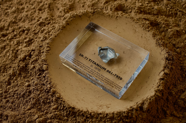 Close-up piece of SR-71 Blackbird titanium embedded in clear acrylic. The fragment is metallic and jagged, which highlights its history from the high-speed reconnaissance aircraft.