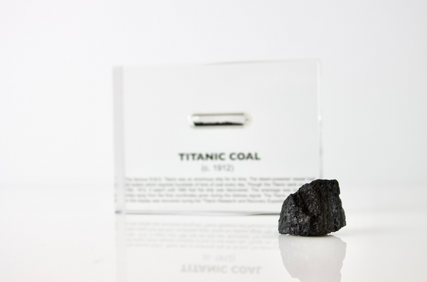 Close-up pieces of coal from the Titanic, contained in a small glass ampule and embedded in clear acrylic. The coal is dark and granular, offering a tangible link to the historic RMS Titanic.