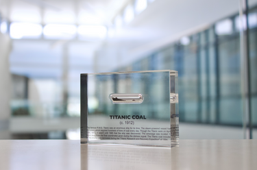 Close-up pieces of coal from the Titanic, contained in a small glass ampule and embedded in clear acrylic. The coal is dark and granular, offering a tangible link to the historic RMS Titanic.
