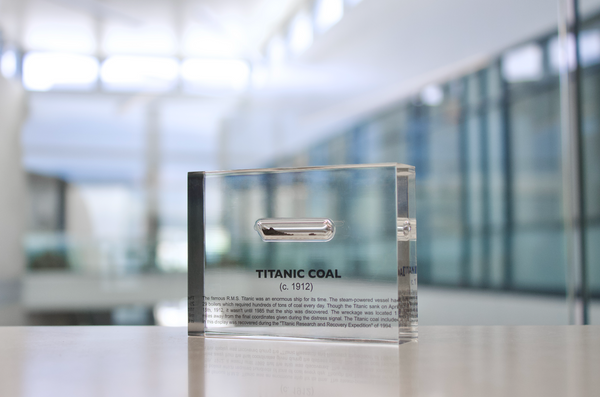 Close-up pieces of coal from the Titanic, contained in a small glass ampule and embedded in clear acrylic. The coal is dark and granular, offering a tangible link to the historic RMS Titanic.