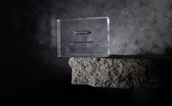 Close-up pieces of coal from the Titanic, contained in a small glass ampule and embedded in clear acrylic. The coal is dark and granular, offering a tangible link to the historic RMS Titanic.