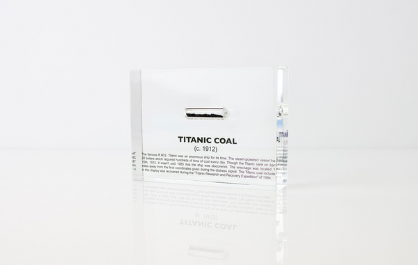 Close-up pieces of coal from the Titanic, contained in a small glass ampule and embedded in clear acrylic. The coal is dark and granular, offering a tangible link to the historic RMS Titanic.