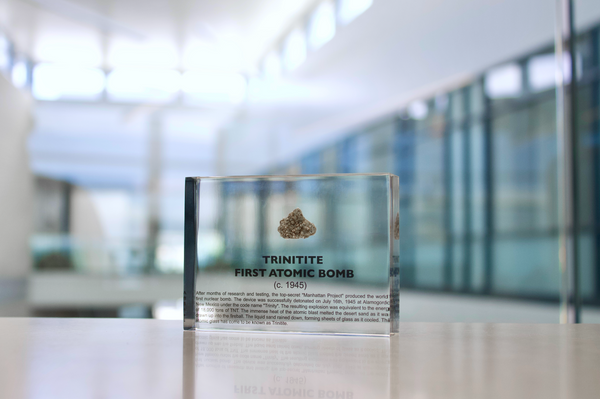 Close-up of a fragment of Trinitite, a material formed from the first atomic bomb explosion, embedded in clear acrylic. The piece has a greenish tint and a rough, uneven surface with visible texture, reflecting its unique origin from the historic Trinity test.