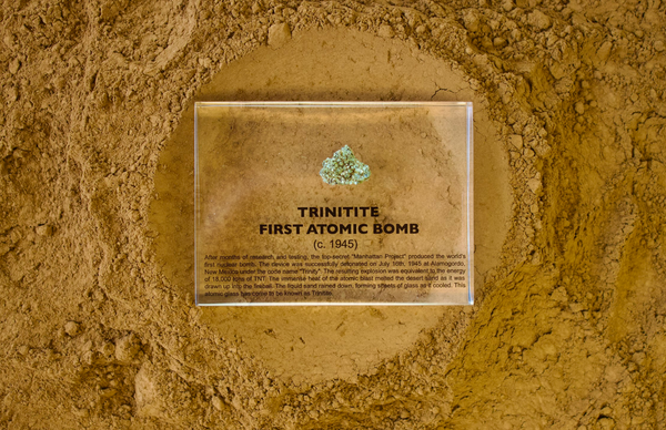 Close-up of a fragment of Trinitite, a material formed from the first atomic bomb explosion, embedded in clear acrylic. The piece has a greenish tint and a rough, uneven surface with visible texture, reflecting its unique origin from the historic Trinity test.