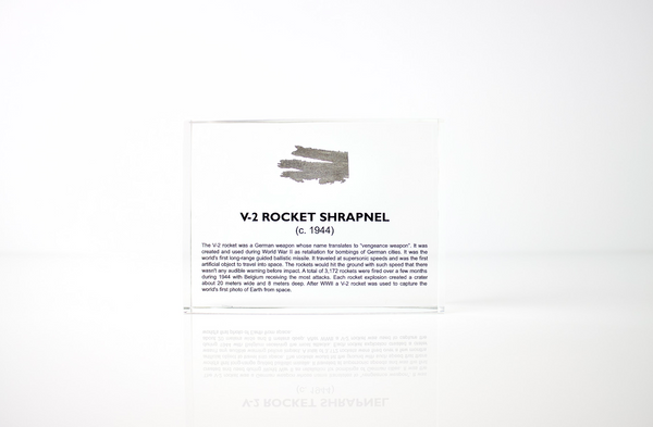 Close-up piece of V-2 rocket shrapnel embedded in clear acrylic. The fragment is metallic and jagged, with a distinctive shape that highlights its history from World War II rocket warfare.