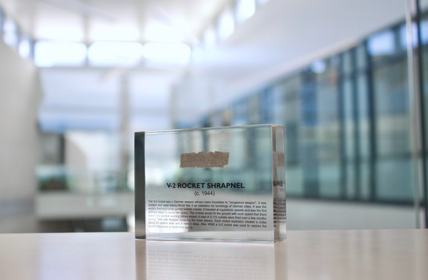 Close-up piece of V-2 rocket shrapnel embedded in clear acrylic. The fragment is metallic and jagged, with a distinctive shape that highlights its history from World War II rocket warfare.