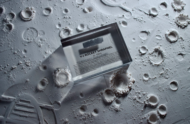 Close-up piece of V-2 rocket shrapnel embedded in clear acrylic. The fragment is metallic and jagged, with a distinctive shape that highlights its history from World War II rocket warfare.