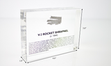 Close-up piece of V-2 rocket shrapnel embedded in clear acrylic. The fragment is metallic and jagged, with a distinctive shape that highlights its history from World War II rocket warfare.