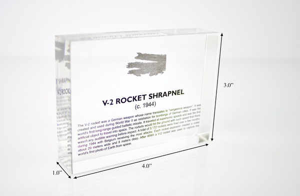 Close-up piece of V-2 rocket shrapnel embedded in clear acrylic. The fragment is metallic and jagged, with a distinctive shape that highlights its history from World War II rocket warfare.