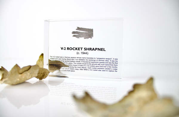 Close-up piece of V-2 rocket shrapnel embedded in clear acrylic. The fragment is metallic and jagged, with a distinctive shape that highlights its history from World War II rocket warfare.