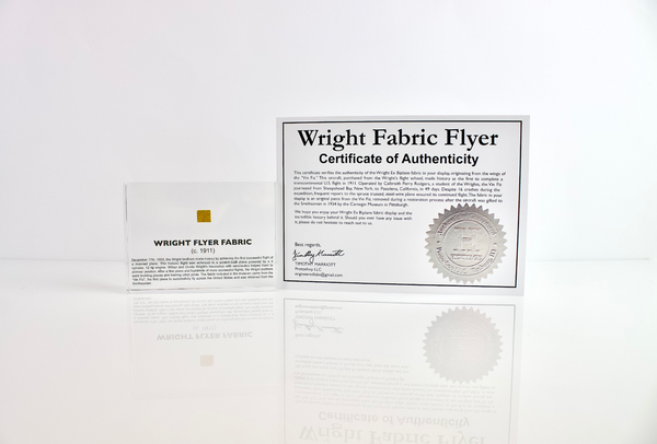 Close-up piece of fabric from the Wright Flyer, encapsulated in clear acrylic. The fabric shows a slightly worn texture with visible stitching, showcasing the early aviation history of the Wright brothers' first successful aircraft.