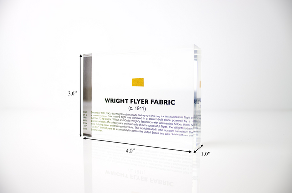 Close-up piece of fabric from the Wright Flyer, encapsulated in clear acrylic. The fabric shows a slightly worn texture with visible stitching, showcasing the early aviation history of the Wright brothers' first successful aircraft.