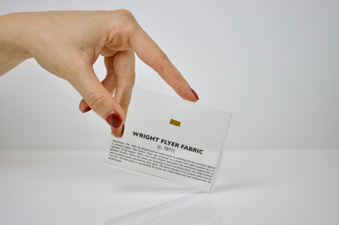 Close-up piece of fabric from the Wright Flyer, encapsulated in clear acrylic. The fabric shows a slightly worn texture with visible stitching, showcasing the early aviation history of the Wright brothers' first successful aircraft.