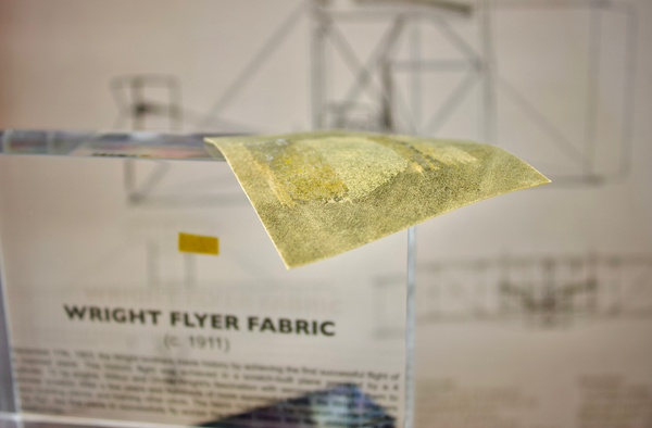 Close-up of a piece of fabric from the Wright Flyer, encapsulated in clear acrylic. The fabric shows a slightly worn texture with visible stitching, showcasing the early aviation history of the Wright brothers' first successful aircraft.