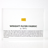 Close-up piece of fabric from the Wright Flyer, encapsulated in clear acrylic. The fabric shows a slightly worn texture with visible stitching, showcasing the early aviation history of the Wright brothers' first successful aircraft.