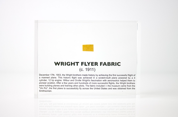 Close-up piece of fabric from the Wright Flyer, encapsulated in clear acrylic. The fabric shows a slightly worn texture with visible stitching, showcasing the early aviation history of the Wright brothers' first successful aircraft.